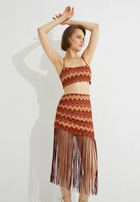 Zweiteiliges Outfit bestehend aus einem crop top mit Zickzack-Muster und einem Rock mit Fransen, in warmen Farbtönen von Rot, Orange und Gold. Das Material scheint strukturiert zu sein.