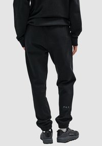 Schwarze Jogginghose mit entspannter Passform, gerippten Bündchen und dem aufgenähten Schriftzug "F.U.T." an dem linken Knöchel. Kombiniert mit dunklen Sportschuhen.