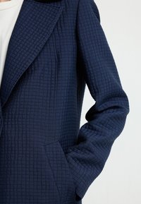 Cappotto strutturato blu navy con motivo trapuntato, ampi rever, maniche lunghe e tasca laterale nascosta. Finissaggio in tessuto liscio.