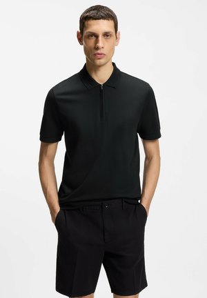 Hombre con camisa polo negra de manga corta con cremallera y pantalones cortos negros, de pie con las manos en los bolsillos contra un fondo liso.