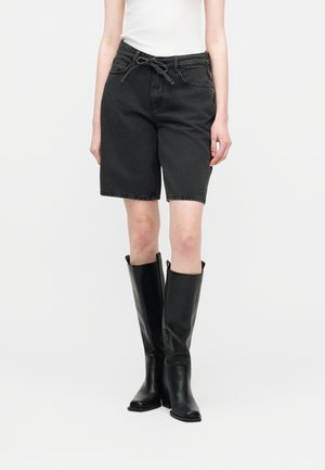 Person iført sorte denimbukser til knæet med snøre i taljen, hvid top og sorte knælange læderstøvler, stående mod en hvid baggrund.