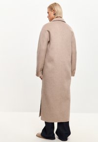 Lungo cappotto in misto lana beige con colletto a revers, aperture laterali e maniche rilassate. Abbinato a jeans a gamba larga scuri e scarpe beige.