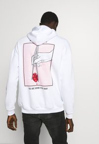White hoodie featuring a graphic on the back: a hand holding scissors above a red heart, with the text "YOU ARE GOING TO BE OKAY."