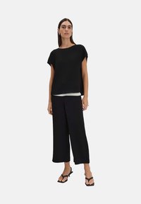 Zwarte textuurtop met korte mouwen, over een witte blouse, gecombineerd met wijde zwarte broek en zwarte sandalen. Minimalistisch ontwerp.