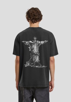 Osoba o kręconych włosach w czarnej koszulce z białą grafiką w stylu szkicu przedstawiającą posąg Chrystusa Odkupiciela na plecach.