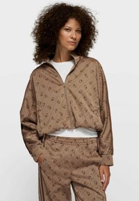 Chaqueta marrón con cremallera y pantalones a juego con un patrón geométrico. Hechos de tela suave con un ajuste holgado y bolsillos laterales.