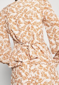 Blumenumhangbluse mit langen Ärmeln, auf weißem Hintergrund mit orangefarbenem Blättermuster. Der gebundene Gürtel betont die Silhouette und hat ein Peplum-Detail.
