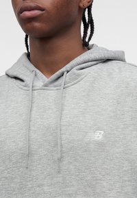 Person wearing a light gray New Balance hoodie with drawstrings, showing shoulder, neck, and braided hair against a plain background.