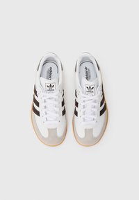 Witte sportsneakers met grijze en bruine suède accenten, voorzien van drie donkere strepen, witte veters en een textuurrijke gumzool.