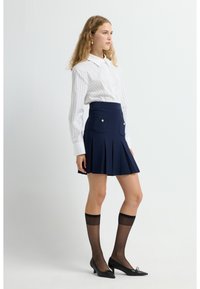 Falda plisada azul marino con dos bolsillos frontales y detalles de botones dorados, combinada con una camisa blanca a rayas y medias negras.