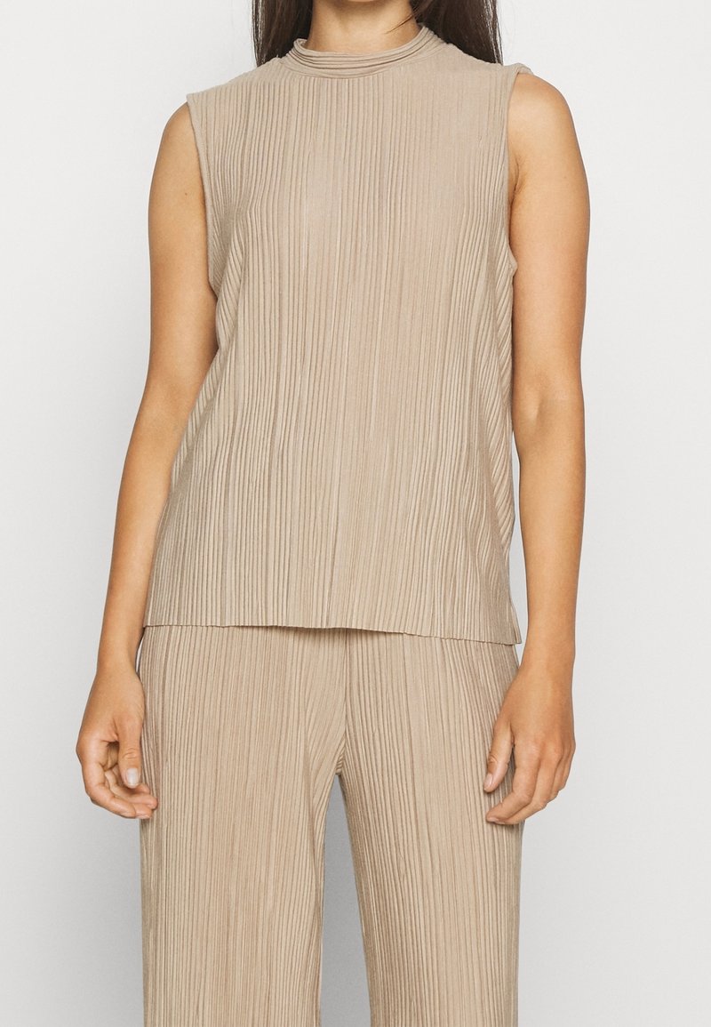 Haut beige sans manches avec un col montant, présentant des plis verticaux et une coupe décontractée, associé à un pantalon plissé assorti.