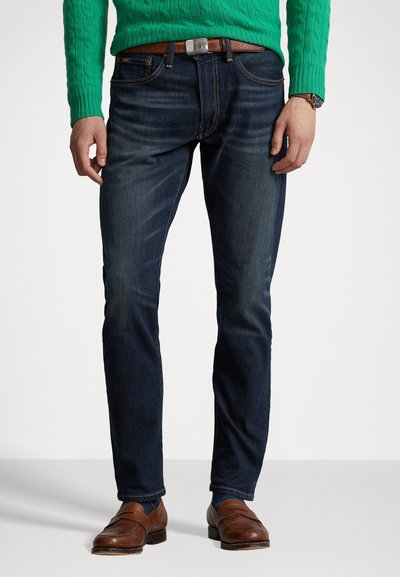 Sötétkék denim farmernadrág, karcsú szabással, finom kopásokkal, klasszikus zsebekkel és varrási részletekkel, barna bőr cipővel párosítva.