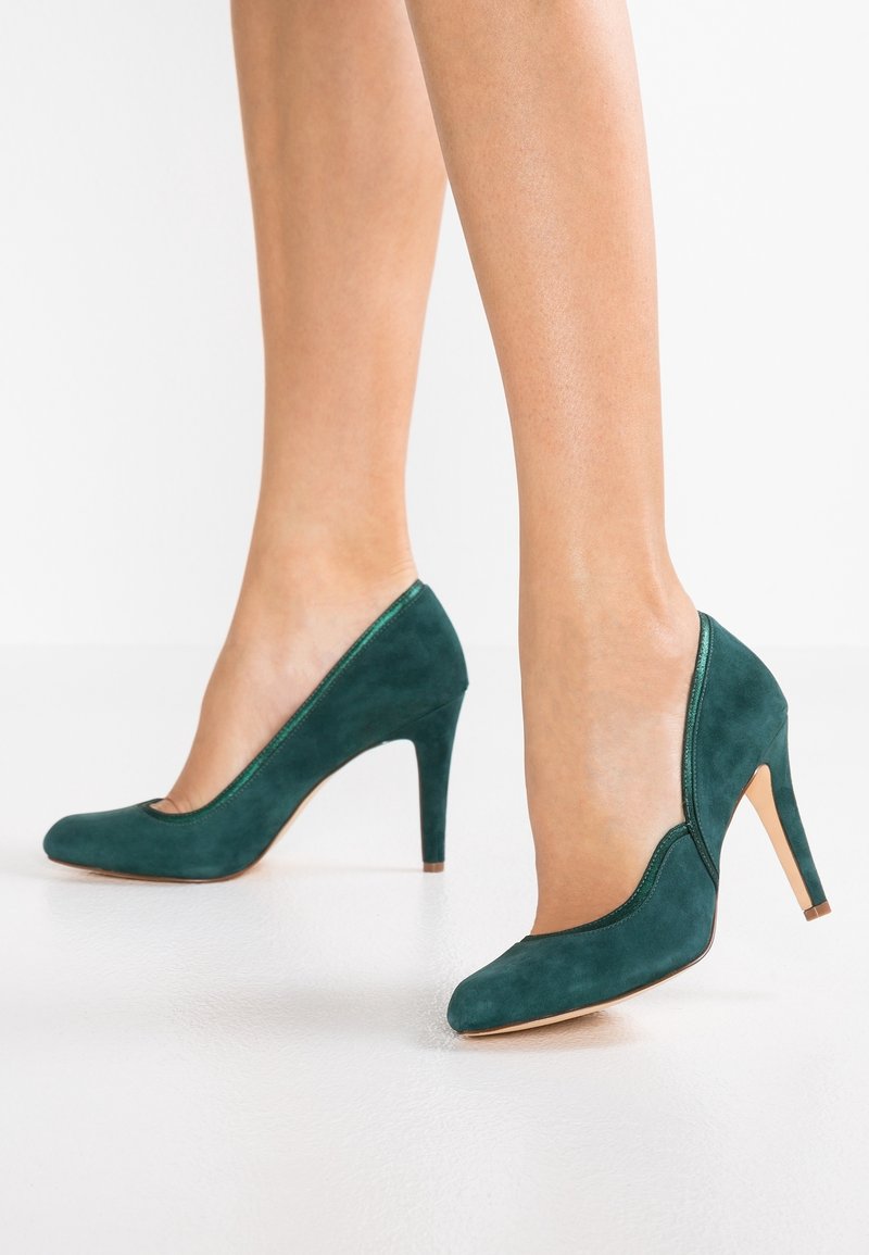 Femme portant des chaussures à talons hauts en daim vert foncé, debout sur un sol blanc avec les mollets et les chevilles visibles.