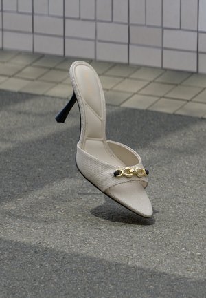Mule beige con tacco alto e dettaglio a catena dorata che si regge in equilibrio sul marciapiede, con parete piastrellata sullo sfondo. Nessuna persona presente.