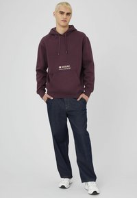 Jeune homme aux cheveux décolorés portant un sweat à capuche Kodak violet, un jean bleu foncé et des baskets blanches, se tenant les mains dans les poches.