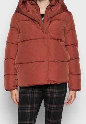 Roestkleurige pufferjas met capuchon, gedragen over zwarte geruite broek, gemodelleerd door een persoon met ontspannen handen langs de zij.