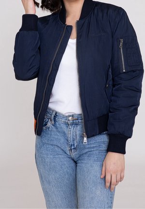 Person wearing a navy blue bomber jacket unzipped over a white ribbed tank top paired with high-waisted light blue jeans.