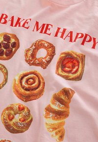 Jasnoróżowa koszulka z ilustracjami różnych wypieków, w tym rogalików, bułeczek cynamonowych i pączków, z napisem "BAKE ME HAPPY" w kolorze czerwonym.