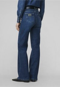 Mørkeblå flarejeans laget av denim, med en lapp med merkevarenavnet på baklommen og en glatt tekstur.