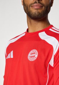 Red FC Bayern München jersey with white accents and three stripes on the shoulder. Features a circular team logo on the chest. Soft fabric.