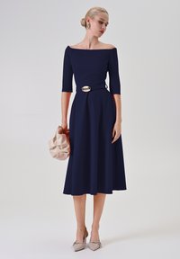 Marineblaues Off-Shoulder-Kleid mit drei Viertel-Ärmeln, figurbetontem Taillenbereich und einer goldenen Schnalle. Begleitet von einer strukturierten beigen Clutch und nudefarbenen High Heels.