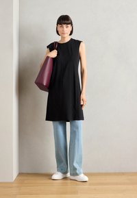 Donna che indossa una tunica nera senza maniche, jeans azzurri chiari, sneakers bianche, mentre tiene una grande borsa tote color mogano, in piedi contro un muro semplice e un pavimento in legno.