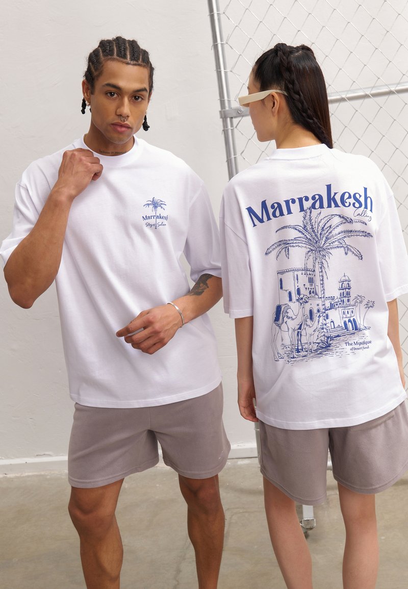 Hvite oversized t-skjorter med blå grafikk av palmer og arkitektur, paret med lysegrå shorts. S Smooth bomullsstoff, løs passform.
