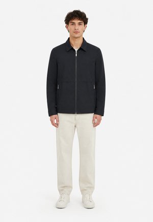Homme portant une veste noire zippée, un pantalon couleur crème et des baskets blanches, debout sur un fond blanc.