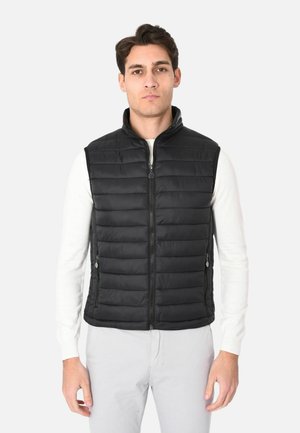 Uomo che indossa un gilet nero trapuntato senza maniche sopra una camicia bianca a maniche lunghe e pantaloni chiari, in piedi davanti a uno sfondo semplice.