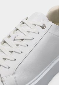 Λευκό δερμάτινο αθλητικό παπούτσι με λευκά κορδόνια, ασημένιες οπές κορδονιών και εμφανείς ραφές, εμφανίζεται σε κοντινή γωνία με φόντο απλό.