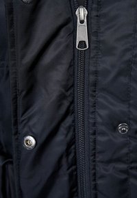 Svart nylonjacka med en central dragkedja, tryckknappar och en slät yta med subtil lyster.