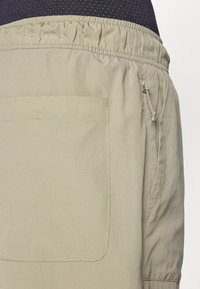 Svijetlosmeđi cargo šorts izrađen od teksturiranog materijala. Ima elastičnu waistband, bočnu džep s patentnim zatvaračem i veliki stražnji džep.