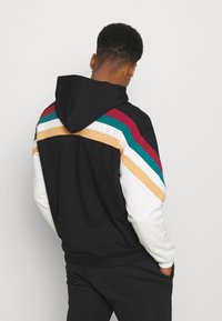 Uomo con capelli ricci corti che indossa un hoodie nero con strisce rosse, verdi, bianche e gialle sulla schiena e maniche bianche, di spalle con le mani in tasca.