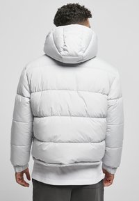 Lys grå pufferjakke med hætte, quiltet design, elastiske manchetter og søm. Stoffet ser glat og isoleret ud.