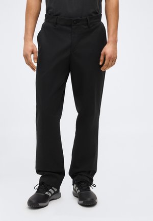 Mees, kes kannab musti sirge säärega pükse ja musti spordijalatsid, seisab valge tausta ees.