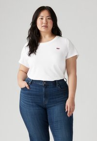Camiseta blanca de algodón con mangas cortas y cuello redondo, con un pequeño logo rojo de Levi's. Combinada con unos pantalones vaqueros de tiro alto en azul oscuro.