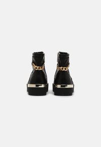 Botines negros de tobillo con un acento de cadena, que presentan un detalle de hardware en tono dorado brillante y un acabado de cuero texturizado. Diseño de suela gruesa.