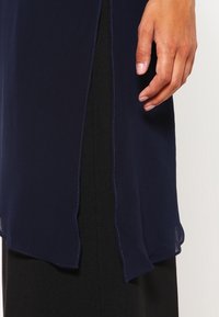 Navy blue flowing fabric top with an asymmetrical hem, featuring a soft texture and visible stitching details. Black fabric underneath.