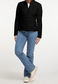 Woman wearing black quilted jacket, blue straight-leg jeans, and white low-top sneakers, standing with legs crossed on a white background.