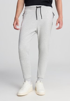 Pantaloni jogger grigio chiaro realizzati in un materiale morbido e elastico. Presentano un elastico in vita con cordino nero, tasche laterali con zip e polsini aderenti.
