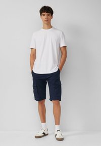 Biała koszulka, granatowe szorty cargo z bocznymi kieszeniami oraz białe sneakersy z czarnymi akcentami. Prosty design, luźny krój, wygodne materiały.
