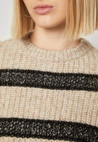 Beige stickad tröja med horisontella svarta ränder och en texturerad, glänsande svart detalj i mitten, med en ribbad hals.