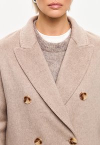 Cappotto doppiopetto in misto lana beige con grandi bottoni lucidi, revers a punta e una texture morbida, indossato sopra un maglione leggero.
