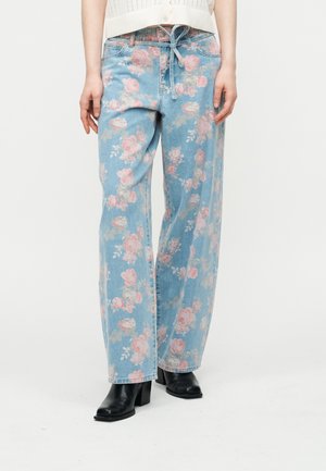 Kobieta w jasnoniebieskich dżinsach z różowym kwiatowym nadrukiem, zestawionych z czarnymi botkami na obcasie i białym krótkim swetrem.