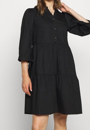 Woman wearing a black, buttoned, tiered dress with puffed 3/4 sleeves, standing against a plain light background.