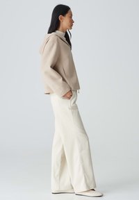 Beige Kapuzenjacke mit lockerem Schnitt, seitlichen Taschen und strukturiertem Stoff, kombiniert mit weiten weißen Hosen. Neutrale Farbpalette.