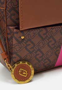 Braune Handtasche mit Muster, die mit einem Lederakzent, goldenen Hardware-Elementen und einem runden Anhänger mit strukturierter Oberfläche und auffälligem Logo-Detail versehen ist.