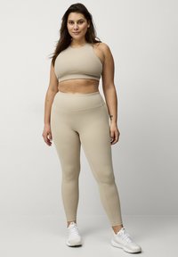 Beige sports-bh og matchende leggings lavet af strækbart materiale. Leggingsene har en høj talje og en glat tekstur, parret med hvide sportssko.