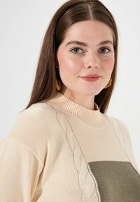 Sweter dziergany z wysokim, prążkowanym kołnierzem, w kolorach kremowym i oliwkowym, z teksturowanymi detalami w sploty na ramionach.