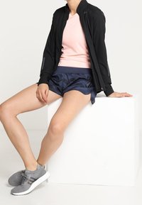 Chaqueta ligera negra, camiseta rosa, pantalones cortos de rayas marino y zapatillas deportivas grises. Diseño elegante con detalles reflectantes y ajuste casual.